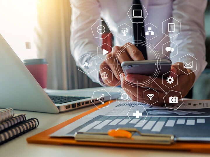 Adult businessman at a desk with an open laptop and charts working on a smartphone with connected devices/programs icons surrounding the phone